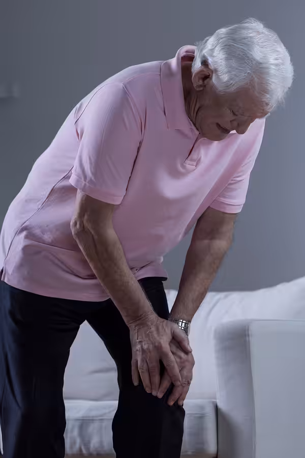 An elderly man with white hair, wearing a pink polo shirt and dark pants, leans forward and clutches his knee in pain while standing next to a white sofa in a dimly lit room.