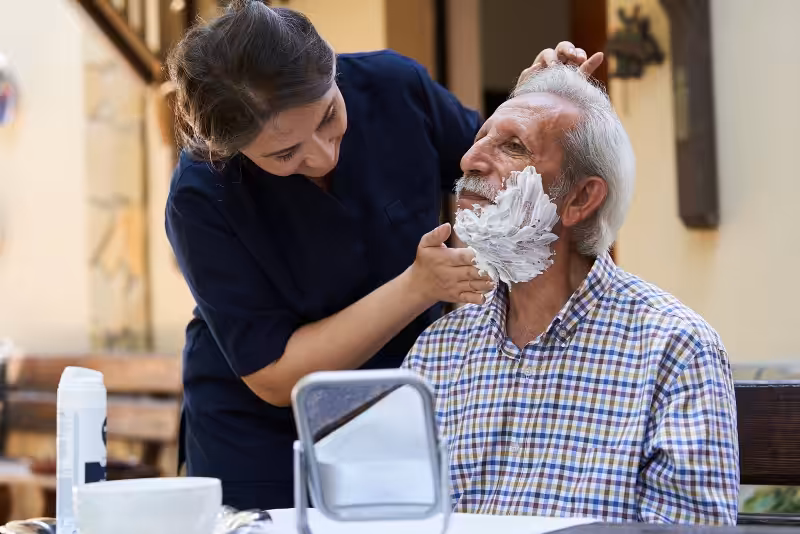 A woman gently applies shaving cream to an elderly mans face as he sits outdoors, smiling and wearing a plaid shirt. A small mirror and shaving supplies are on the table in front of them.