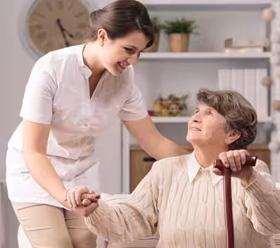 A young caregiver smiles and holds hands with an elderly woman seated with a walking cane, indoors with a clock and shelves in the background.