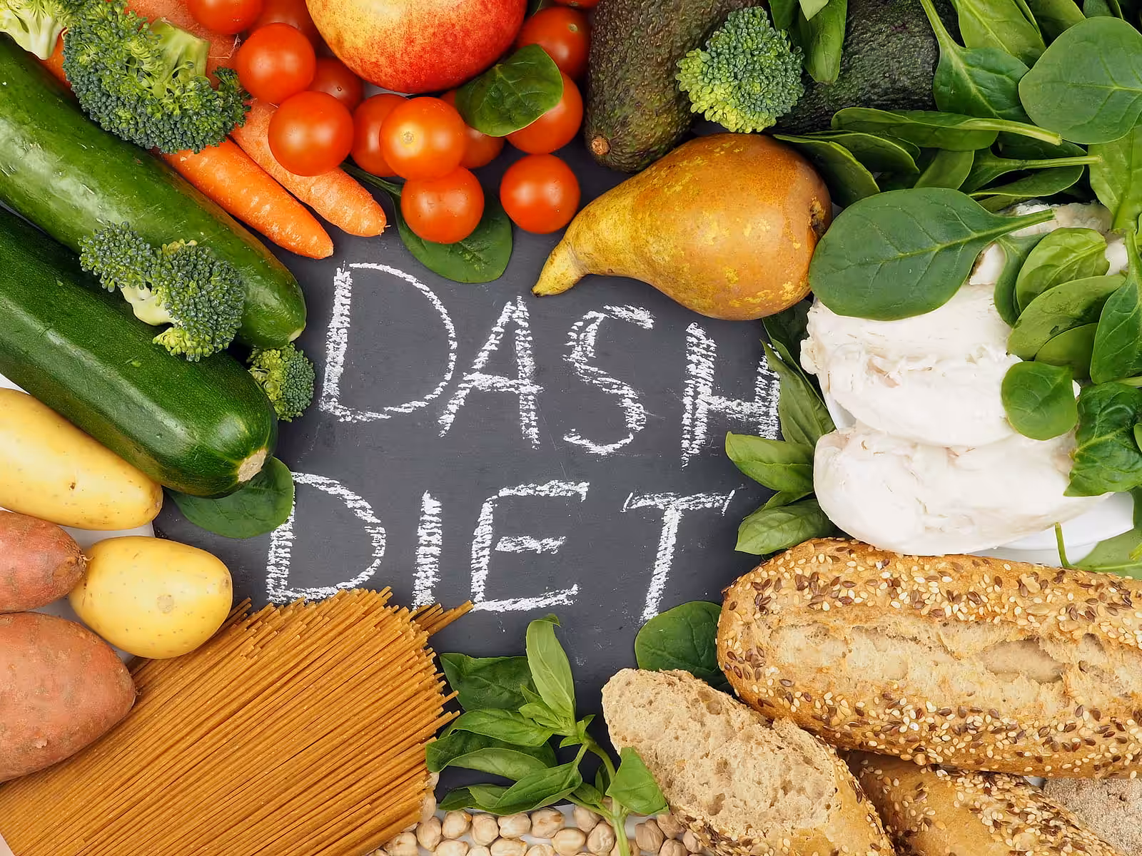 A chalkboard with DASH DIET written on it is surrounded by fresh vegetables, fruits, chicken breast, whole grain bread, pasta, potatoes, beans, and leafy greens.