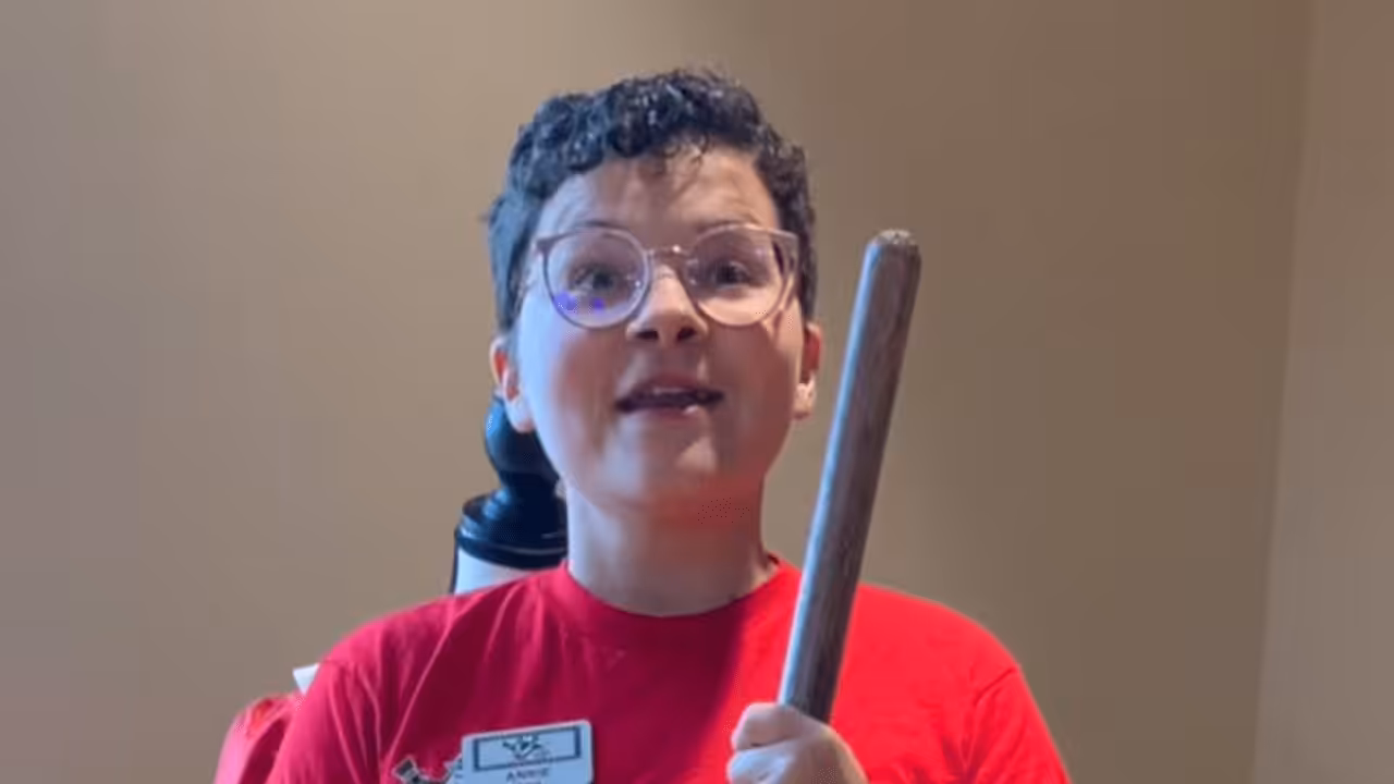 A young person with short curly hair and glasses, wearing a red shirt and name tag, holds a wooden stick and smiles while standing indoors against a beige wall.