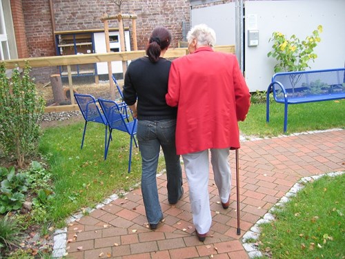 A younger person walks arm-in-arm with an older person using a cane along a brick path in a garden with blue benches and green grass.