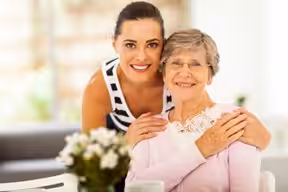 A young woman smiles and hugs an older woman from behind as they sit together indoors. Both are smiling warmly at the camera, and a vase of flowers is on the table in front of them.