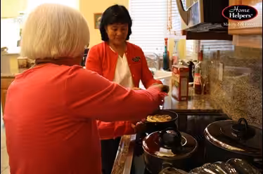 Two women are in a kitchen; one is cooking food in a pan while the other assists and smiles. The Home Helpers logo appears in the top right corner, suggesting a home care or assistance service.