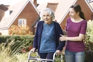 A young woman supports an elderly woman using a walker as they walk outside near houses, both smiling and enjoying a sunny day.