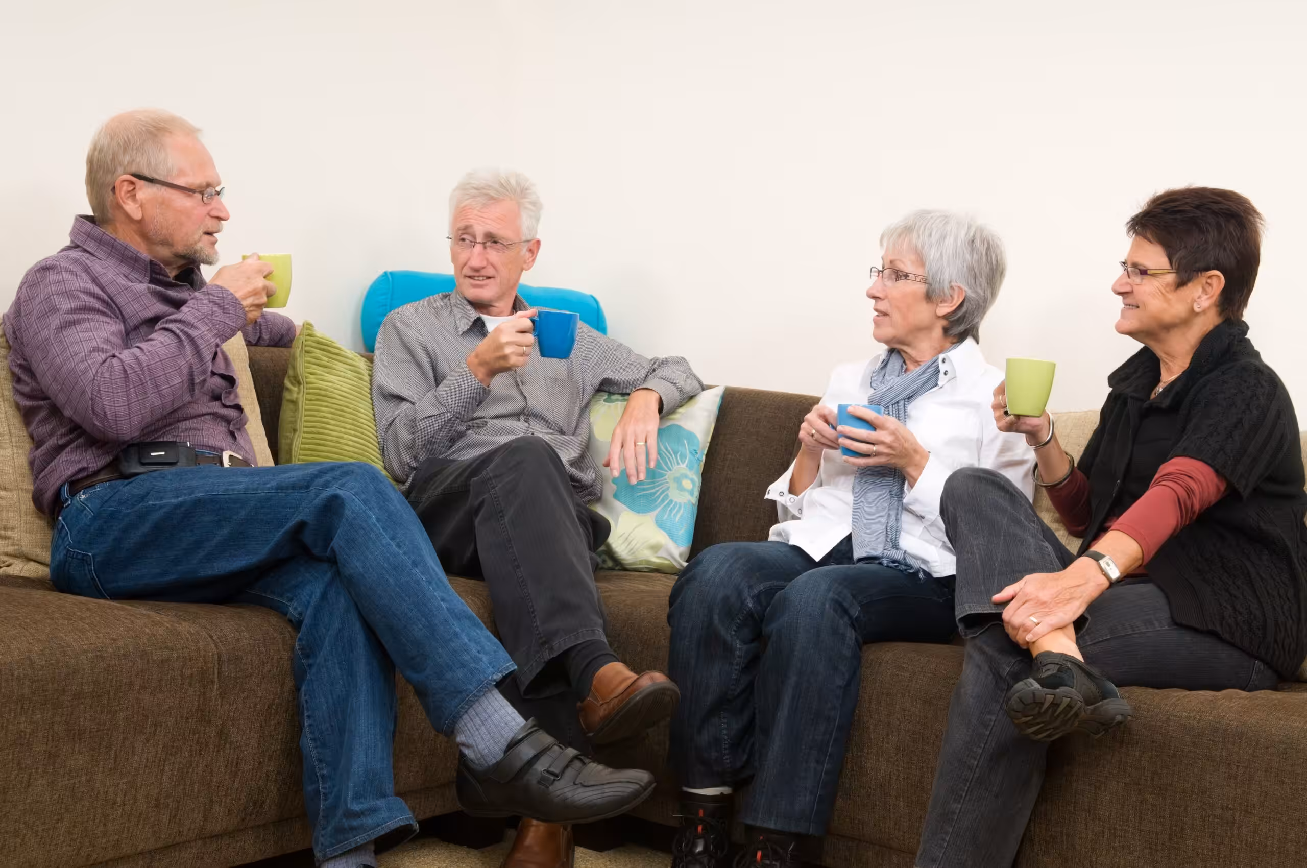 Four older adults sit together on a brown sofa, smiling and drinking from mugs. They appear to be engaged in a friendly conversation in a cozy, relaxed indoor setting.