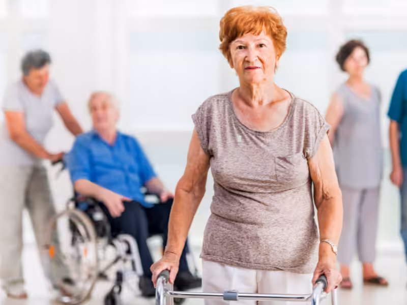 An elderly woman using a walker stands in focus, while other seniors, including a man in a wheelchair and two women, interact in the blurred background of a bright room.