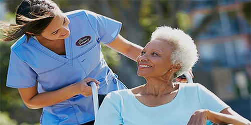 A nurse in blue scrubs smiles and talks with an elderly woman in a wheelchair outdoors, both appearing happy and engaged.