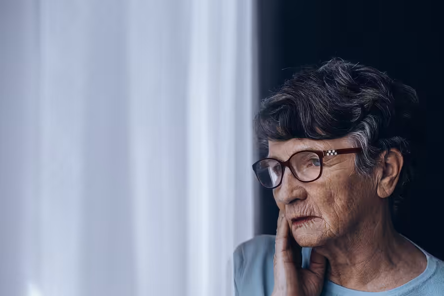 Elderly woman with short gray hair and glasses looks thoughtfully out a window, resting her hand on her cheek, with soft natural light illuminating her face.