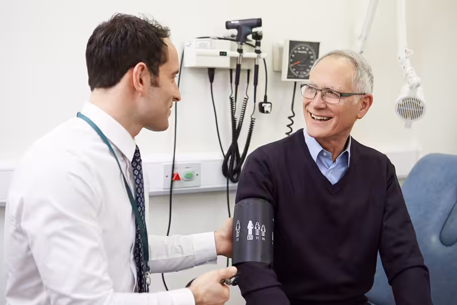 A doctor checks an older mans blood pressure in a medical office. The man is smiling and sitting on an exam table, while the doctor adjusts the blood pressure cuff on his arm. Medical equipment is visible on the wall behind them.