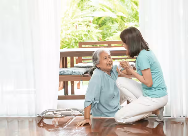 An elderly woman sits on the floor by a walker, smiling as a younger woman kneels beside her, holding her hand to offer support. They are inside a bright room with large windows overlooking greenery.