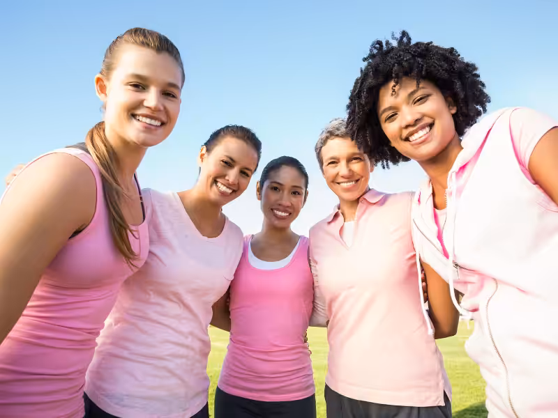 Five women standing close together outdoors, smiling at the camera. They are all wearing pink shirts, suggesting unity or support for a cause, with green grass and blue sky in the background.