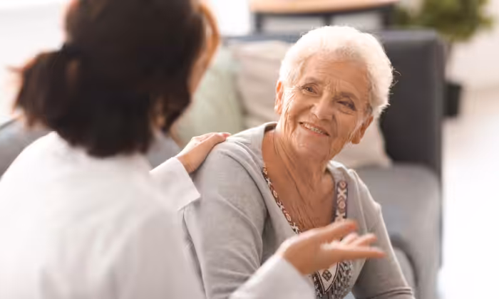 An older woman with short gray hair smiles and talks to another person, who rests a reassuring hand on her shoulder. They are seated indoors on a couch, creating a warm and supportive atmosphere.