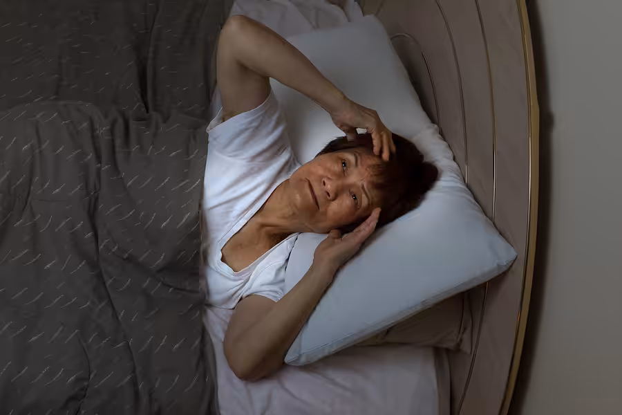 An older adult lies in bed at night, looking up with a concerned expression, one hand resting on their forehead. They are partially covered by a gray blanket and lying on a white pillow.