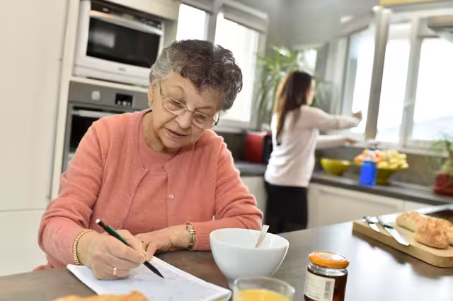 An elderly woman wearing glasses writes in a notebook at a kitchen table with food items, while a younger woman prepares something at the counter in the background.
