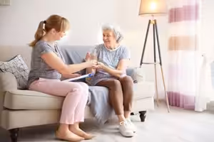 A younger woman and an older woman sit together on a sofa in a bright living room, smiling and having a conversation. The younger woman holds a clipboard and pen. A lamp and curtains are in the background.