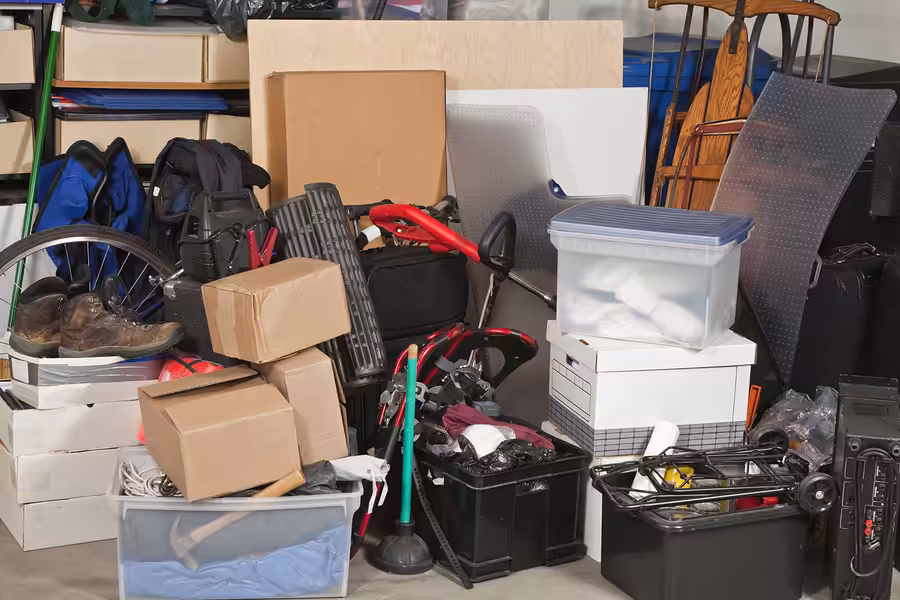 A cluttered storage area filled with boxes, plastic bins, a wheelchair, a pair of shoes, a folding chair, and various household items stacked and piled together.