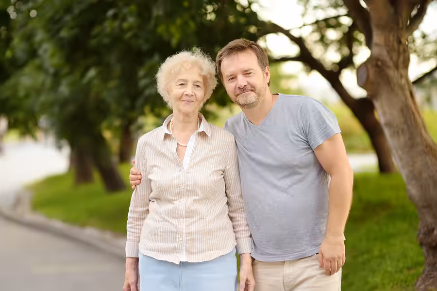An older woman and a middle-aged man stand close together outdoors, smiling at the camera, with trees and greenery in the background. The man has his arm around the womans shoulders.