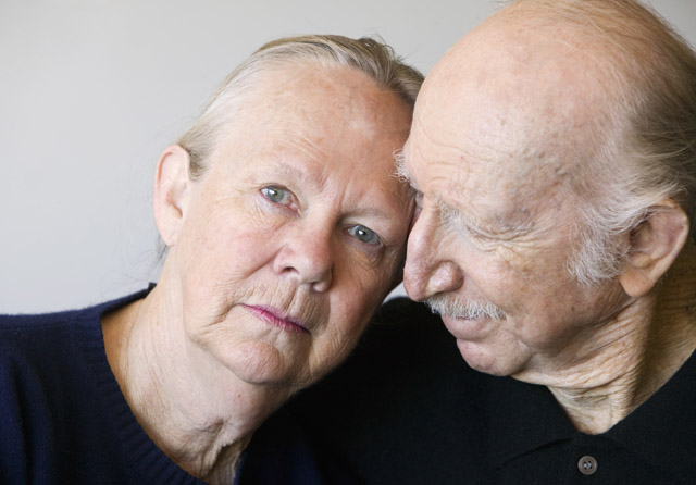 An elderly woman and man sit closely together, the woman looking directly at the camera while the man leans in, touching his forehead to hers in a tender, intimate moment.