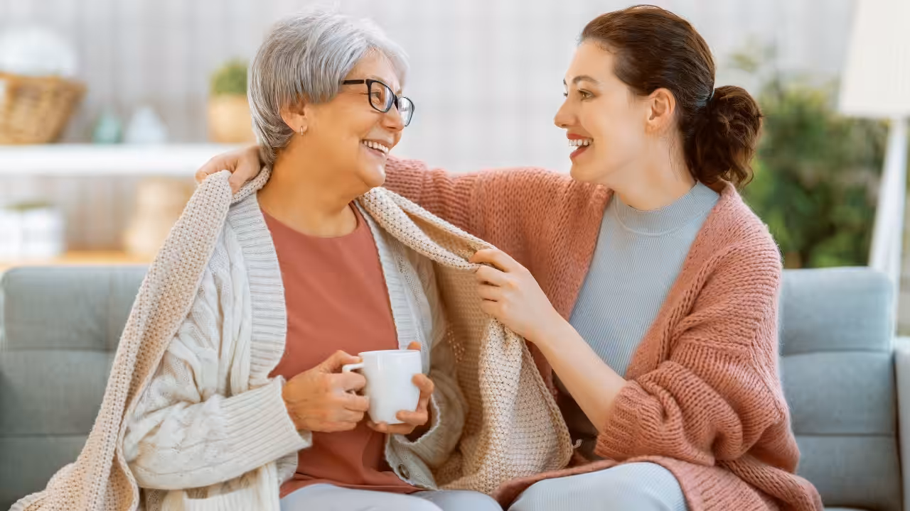 A smiling older woman holding a mug sits beside a younger woman who is wrapping a cozy blanket around her shoulders. Both appear happy, sitting on a couch in a bright, comfortable room.