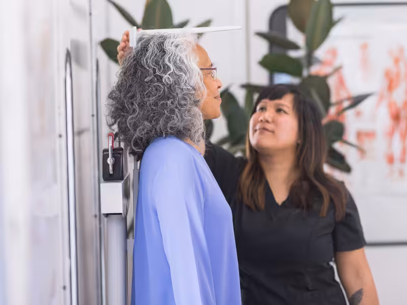 A healthcare professional measures the height of an older woman with gray curly hair using a stadiometer in a medical office.