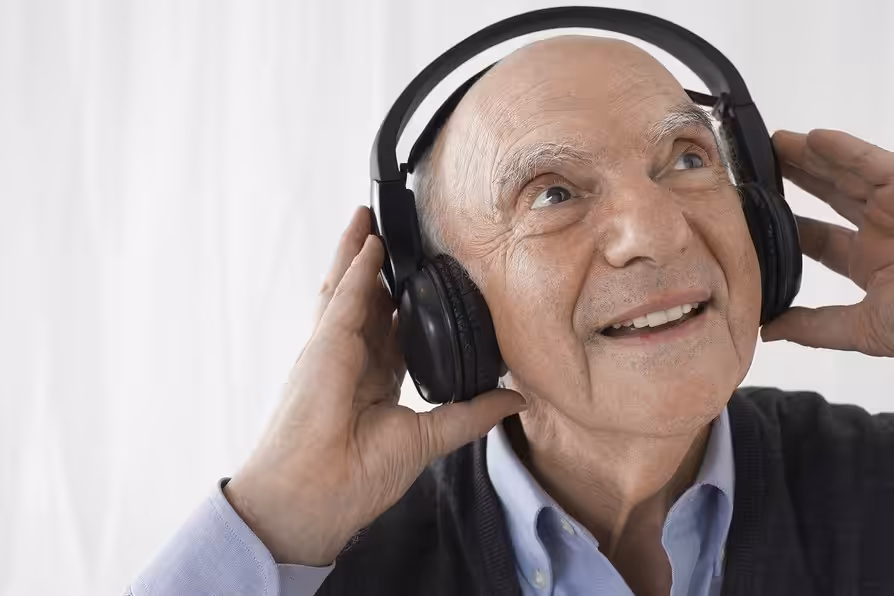 An older man with a bald head smiles and looks upward while enjoying music on large black headphones. He wears a light blue shirt and a dark sweater, with one hand touching the headphones.