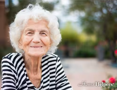 Smiling elderly woman with white hair wearing a black and white striped shirt sits outdoors. The background is blurred with greenery and flowers. Home Helpers logo is in the bottom right corner.