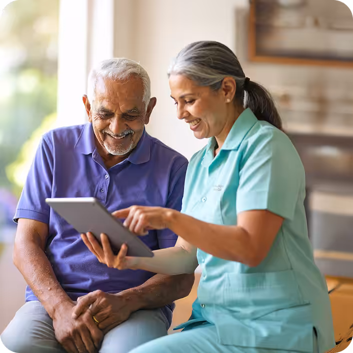 A smiling elderly man sits next to a woman in medical scrubs who is showing him something on a tablet. They both appear engaged and happy in a well-lit room.