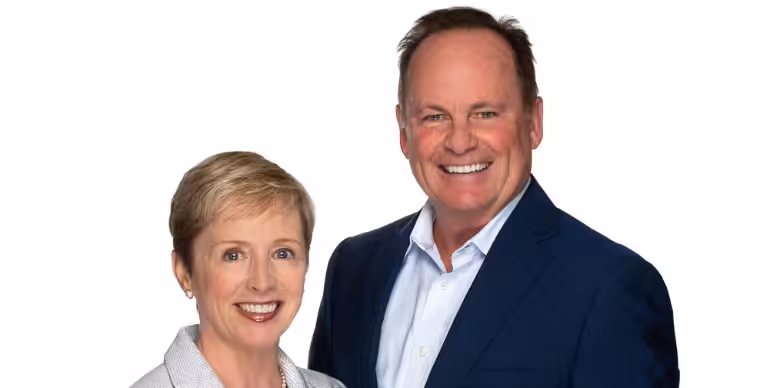 A woman with short blonde hair and a light-colored blazer stands next to a man with short brown hair wearing a navy blue suit and white shirt, both smiling in front of a white background.