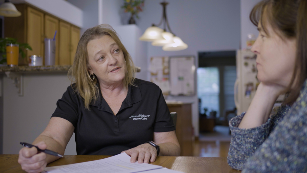 A woman in a black Home Helpers Home Care shirt sits at a table with paperwork, talking to another woman in a patterned shirt in a home kitchen setting.