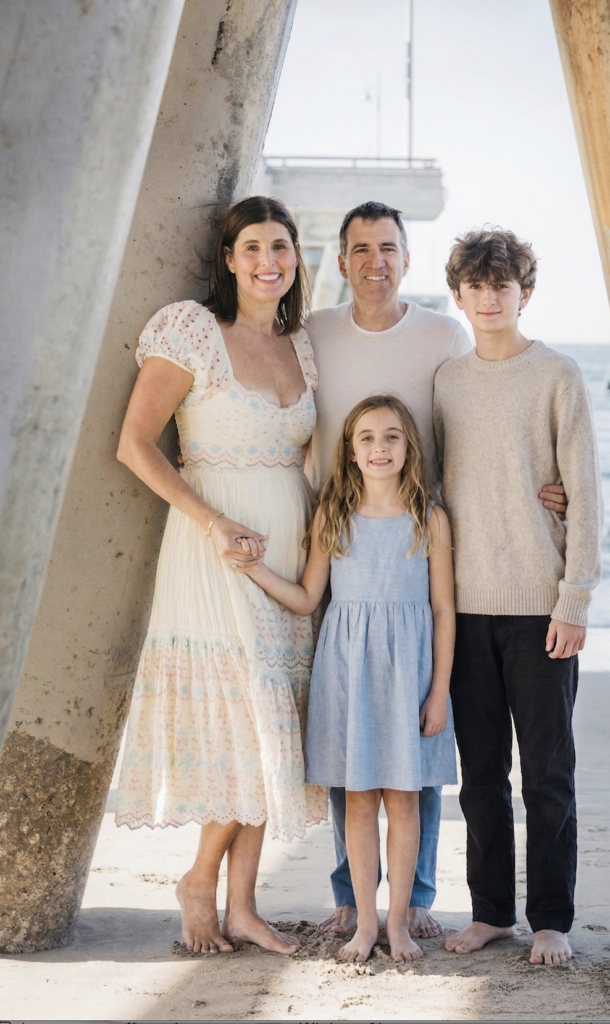 A family of four stands barefoot on a sandy beach under a pier, smiling at the camera. The mother wears a light dress, the father wears a white shirt, and their son and daughter wear a sweater and a blue dress, respectively.