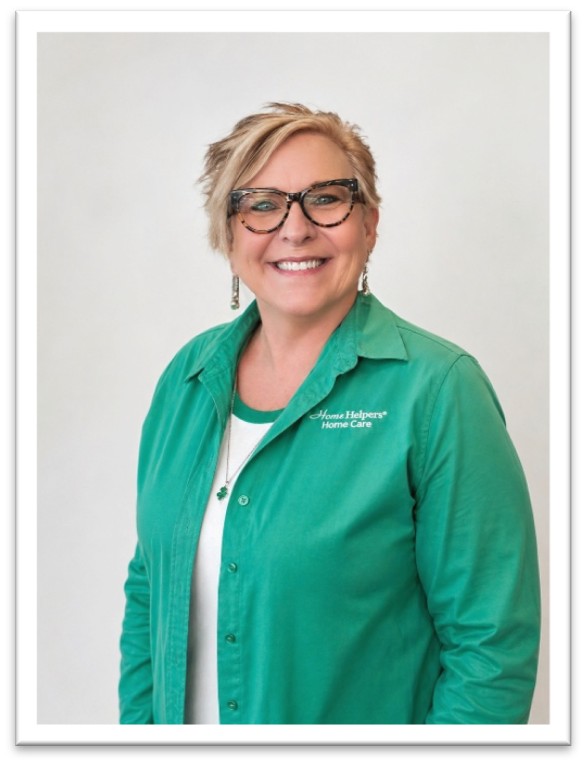 A smiling woman with short blonde hair and glasses wears a green button-up shirt over a white top. The shirt has a “Home Helpers Home Care” logo. She stands against a plain light background.