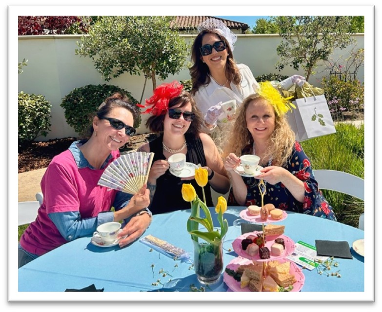 Four women sit at a table outdoors, smiling and holding teacups. The table has a blue cloth, yellow tulips, and a tiered tray with treats. Two women wear fascinators, one holds a fan, and another stands holding a gift bag.