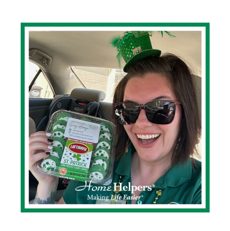 A smiling woman wearing a green mini hat and sunglasses holds a package of green St. Patrick’s Day cookies inside a car. The image has a white border with the text “Home Helpers® Making Life Easier®” at the bottom.