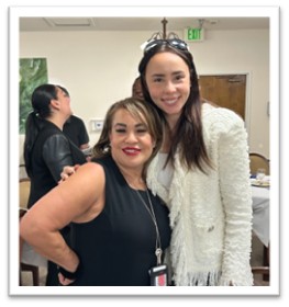 Two women smile and pose together indoors at a social event. One wears a sleeveless black dress and red lipstick; the other wears a white textured jacket and sunglasses on her head. People and tables are visible in the background.