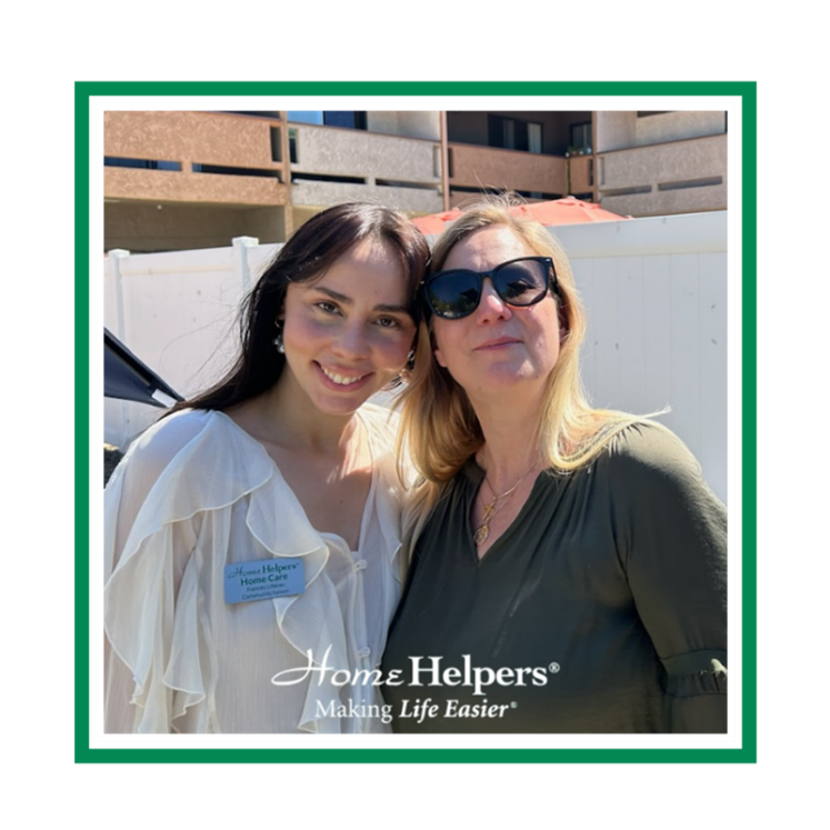 Two women stand close together and smile outdoors. One wears a white blouse with a name tag, the other wears sunglasses and a green blouse. A white fence and buildings are in the background. Home Helpers® Making Life Easier® is at the bottom.