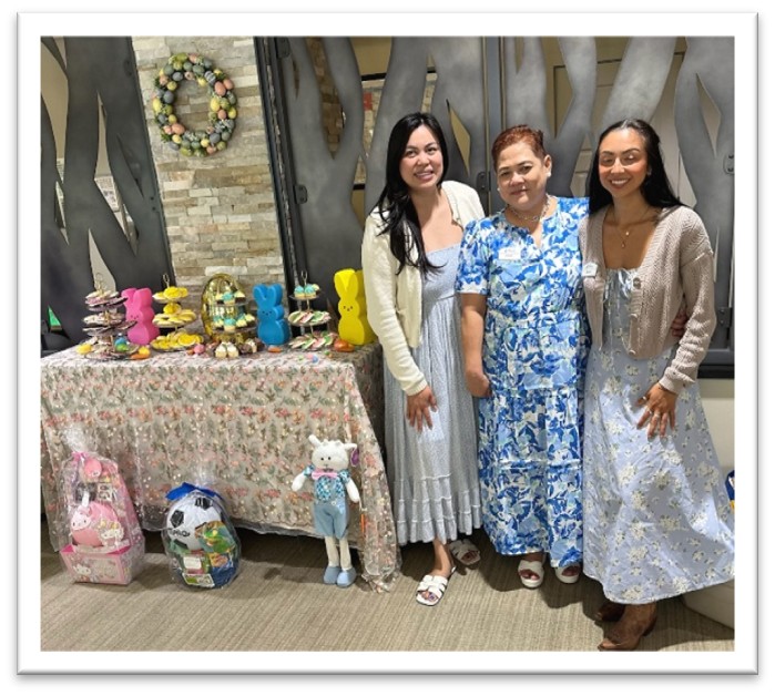Three women in floral dresses stand by a decorated table with Easter-themed treats, colorful bunnies, and a stuffed bunny, with a festive wreath hanging on the wall in the background.