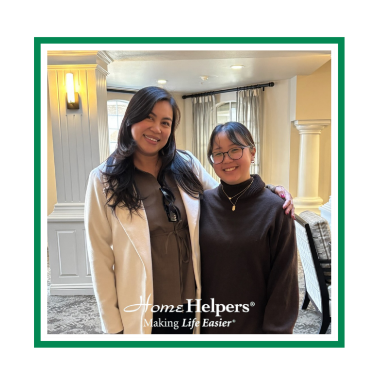 Two women stand indoors, smiling at the camera. One wears a white blazer, the other a brown sweater. They appear happy and close. The image has a green border and the text, Home Helpers® Making Life Easier® at the bottom.