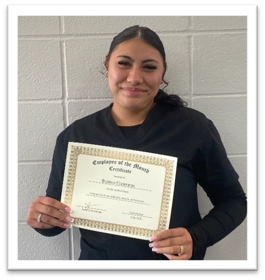 A woman in black scrubs stands smiling, holding an Employee of the Month certificate in front of a light gray brick wall.