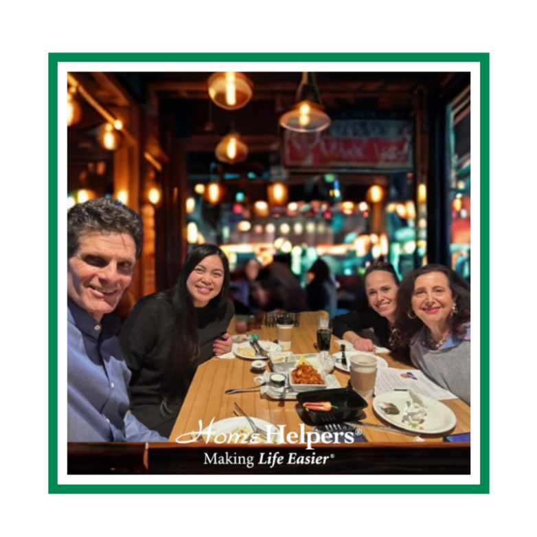 Four people smiling and sitting at a restaurant table with food and drinks. The image has a Home Helpers® Making Life Easier® logo at the bottom and is framed by a green border. The background has warm, ambient lighting.