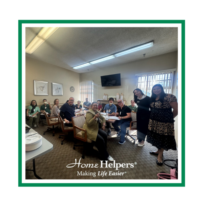 A group of people sit and stand around a table in an office, smiling at the camera. The image is framed with the Home Helpers logo and the slogan Making Life Easier.