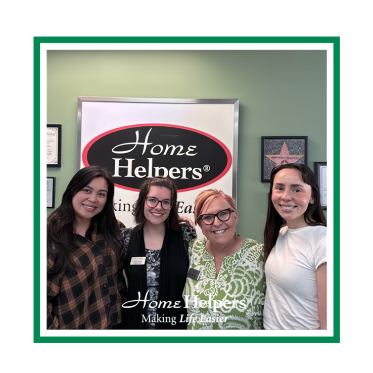 Four women stand smiling in front of a Home Helpers sign in an office setting. They are close together, wearing casual and business-casual clothing. The Home Helpers logo and slogan appear at the bottom of the image.