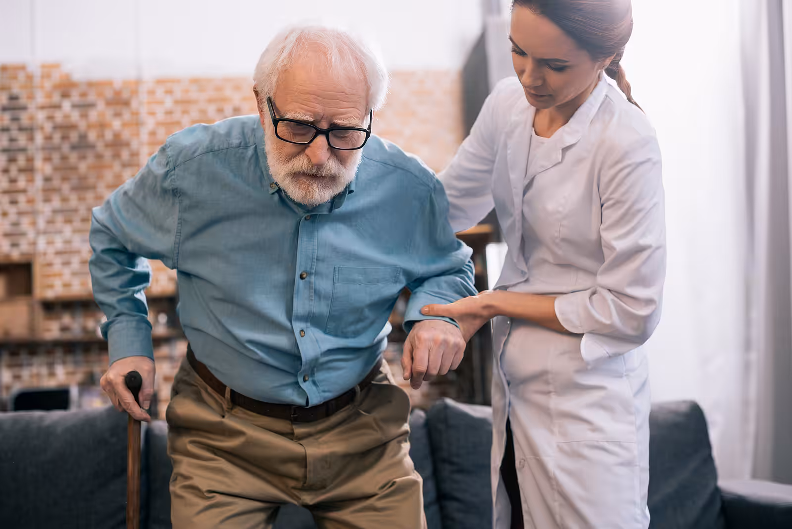 A young female caregiver in a white coat helps an elderly man with glasses and a cane stand up from a sofa, supporting him by the arm in a home setting.