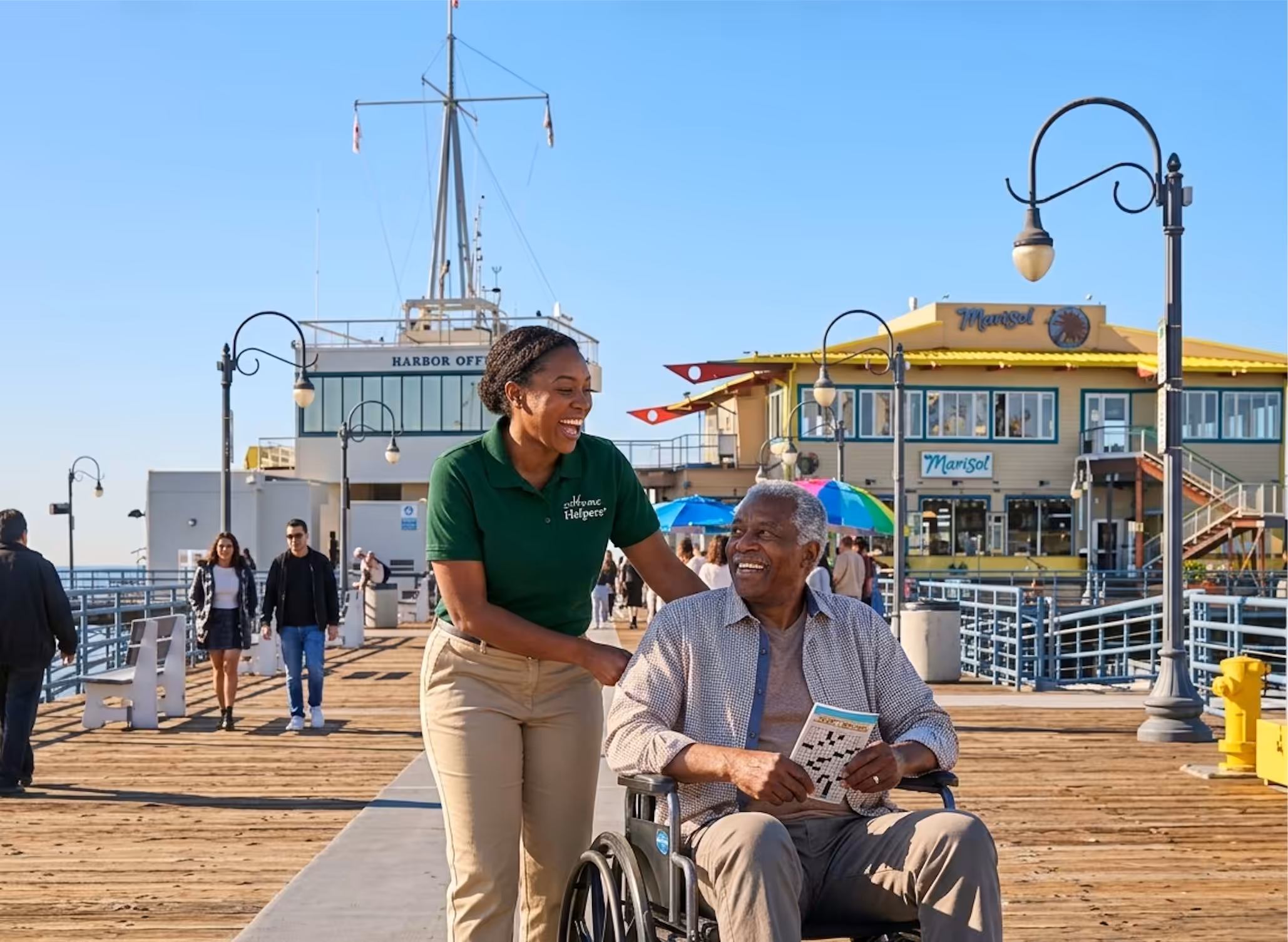 A smiling woman pushes an elderly man in a wheelchair along a sunlit pier, with shops, people walking, and colorful umbrellas in the background. Both appear happy and are enjoying the day outdoors.