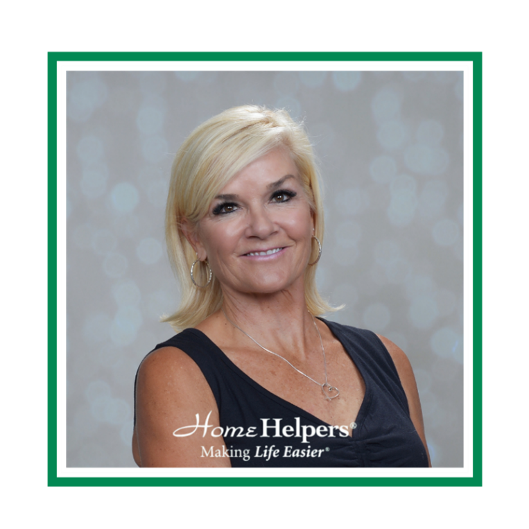 Smiling woman with short blonde hair wearing a sleeveless dark top and hoop earrings, posing in front of a blurred light gray background. Home Helpers® Making Life Easier® logo is at the bottom of the image.