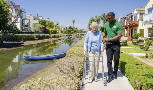 A young man assists an elderly woman using a walker as they walk along a sunny canal-side path, surrounded by greenery, with houses and a canoe visible in the background.