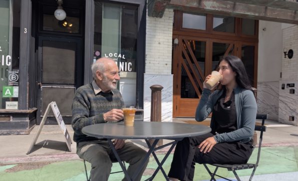 An older man and a younger woman sit at a round outdoor table, enjoying drinks and talking in front of a café with “Local Organic” on the window. The woman drinks from a cup while the man holds his cup, smiling.