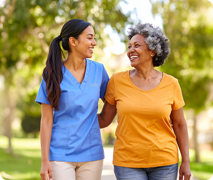A smiling healthcare worker in blue scrubs walks arm-in-arm with an older woman in a yellow shirt outdoors, both looking at each other and appearing happy. Trees and sunlight are visible in the background.