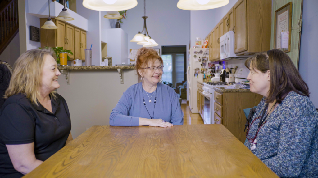 Three women sit around a wooden dining table in a cozy kitchen, smiling and talking with each other. The kitchen is warmly lit with hanging lights and has wooden cabinets and various kitchen appliances.