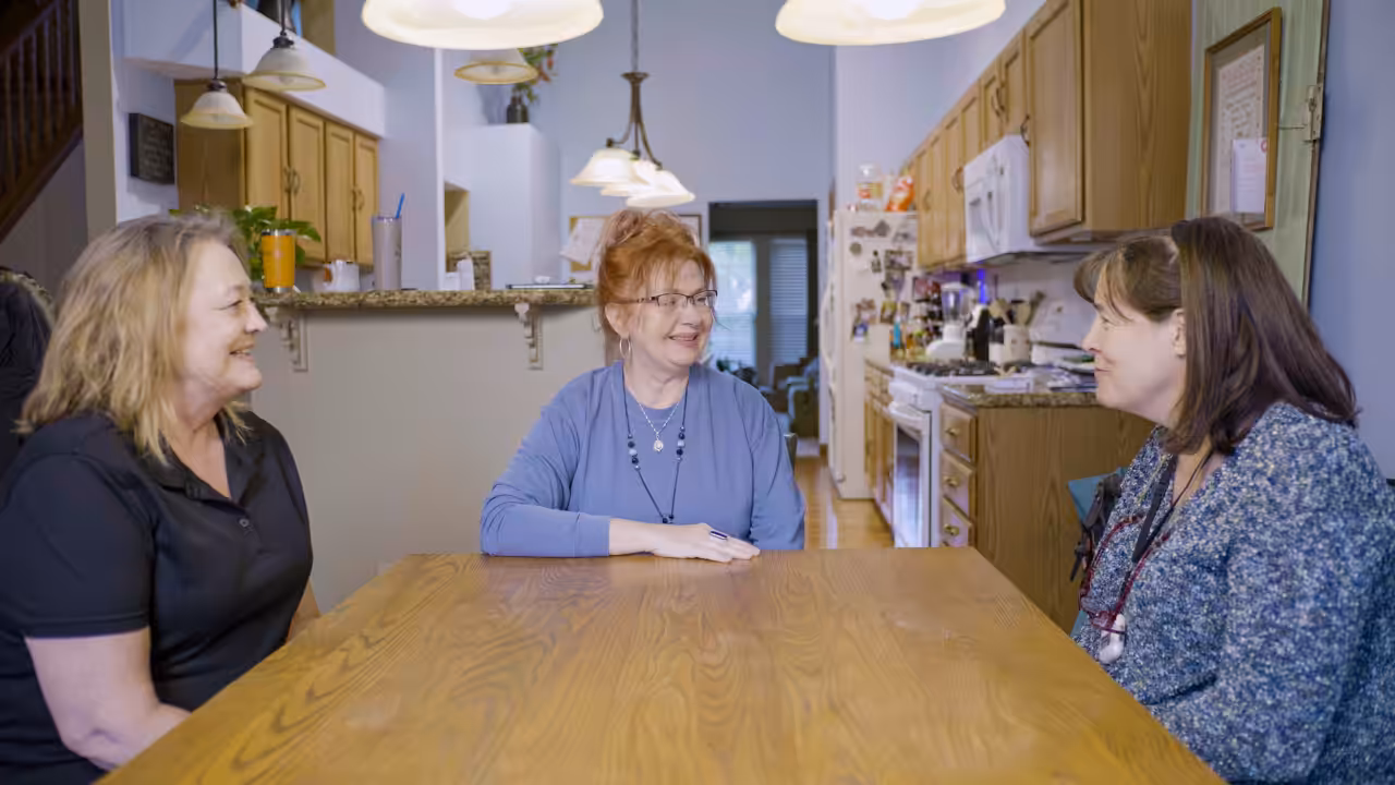 Three women sit around a wooden dining table in a cozy kitchen, smiling and talking with each other. The kitchen is warmly lit with hanging lights and has wooden cabinets and various kitchen appliances.
